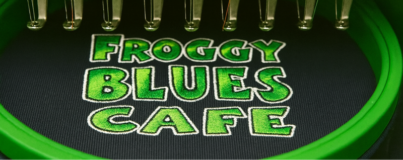 Embroidery machine needle plate with 'Froggy Blues Cafe' text on a dark fabric background